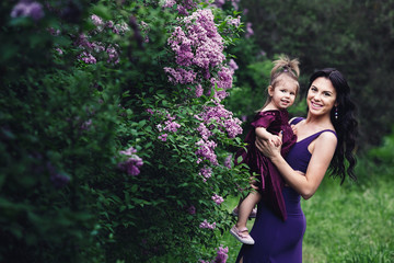 cheerful mother and little daughter in purple dresses playing in nature near the blossoming lilac