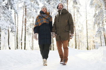 Full length portrait of happy couple holding hands in winter forest and walking towards camera, copy space