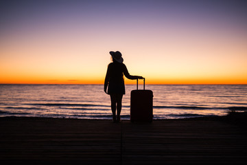 Travel, vacation and summer trip concept - silhouette of young woman in summer dress and hat looking to a sea