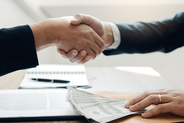 Two corporate businessmen shaking hands while one man places money on document in office room with corruption concept.