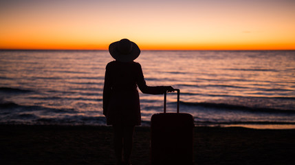 Obraz premium Travel, vacation and holiday concept - Young woman standing with suitcase on sandy beach at sea at the sunset
