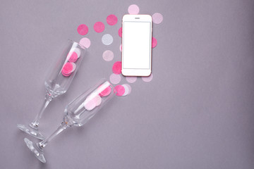 Champagne glasses and mobile phone with pink confetti on gray background