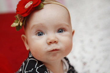 Beautiful five-month toddler girl with a flower on her head dress on a red background. Baby look. Maternal care. Childcare. Close-up.