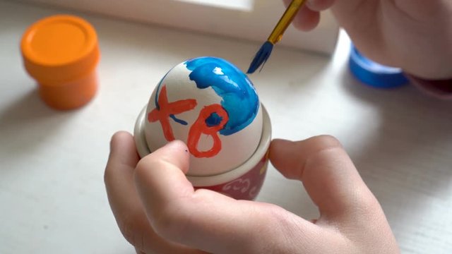 Close-up, the child paints a white Easter egg with a brush and blue paint. The view from the top. 4K. 25 fps