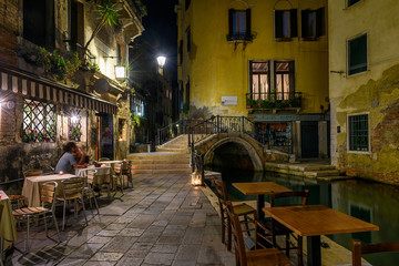 Fototapeta na wymiar Narrow canal with bridge and tables of restaurant in Venice, Italy. Architecture and landmark of Venice. Night cozy cityscape of Venice.