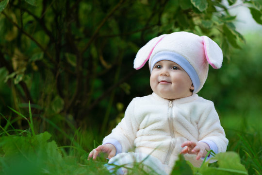 Child girl with chubby cheeks in bunny costume seating on grass in park.