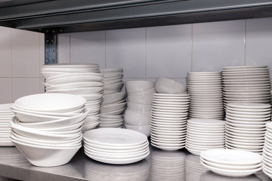 A Pile Of White Clean Ceramic Porcelain Plates On A Metal Rack In The Back Of The Restaurant. Concept Of Preparation For Banquet, Catering, Buffet