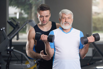 Fototapeta na wymiar Elderly man and trainer posing with dumbbells in gym.