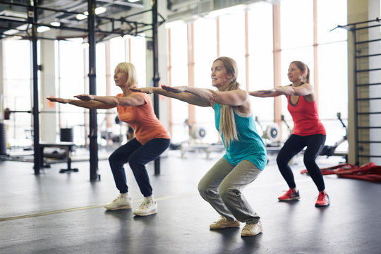 Group of mature active females squatting and stretching arms forwards during workout in fitness center
