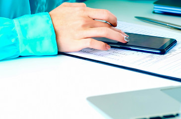 Close-up of female hands using smart phone while working on computer at modern office interior, businesswoman typing text message on her cellphone at loft interior.