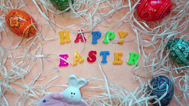 Two plates (pink and blue) with Easter bunnies are placed next to painted multicolored Easter eggs, decorative straw and the words "Happy Easter" written in multicolored letters. Top view