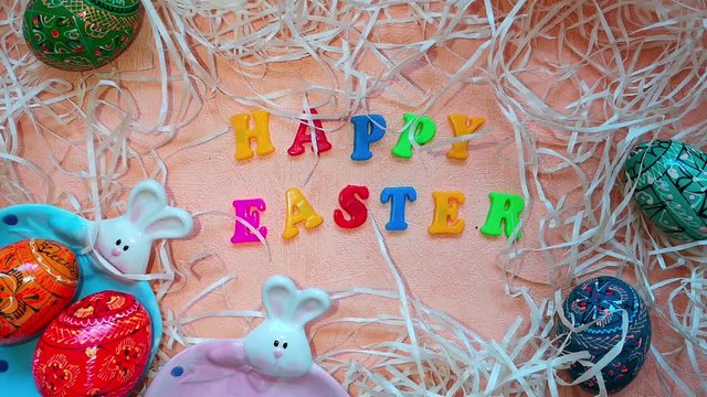 Two painted Easter eggs fall on a plate with Easter bunny, next to it are painted colorful Easter eggs, decorative straw and words "Happy Easter" written in different colored letters. Top view