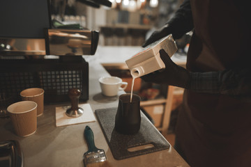 Barista pours milk from a pack of pitcher. Professional barista in the workplace prepares cappuccino. Barista Concept. Cooking coffee with milk in the coffee shop. Workplace Barista.