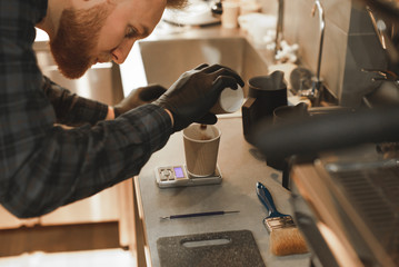 Concentrated barista with a beard is pouring coffee to the paper cup and weighing it on the kitchen scales. Focused bartender weighs coffee on the electronic scales at coffee shop. Close up photo.