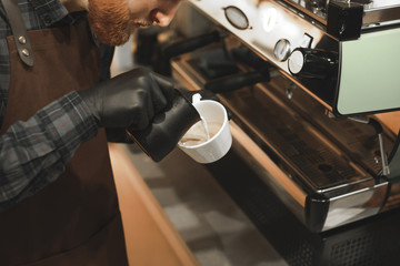 Close up view on barista hands pouring milk and making coffee art on a cappuccino at coffee machine. Background photo of concentrated bartender preparing tasty coffee drink.