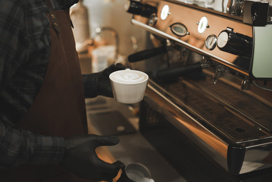 Close up photo of barista in the apron and gloves holding a cup with latte with beautiful latte art. Background photo of bartender at coffee machine with beautiful coffee art.