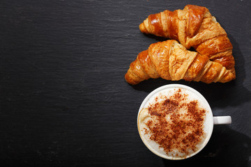 Cup of cappuccino coffee and croissants on dark table