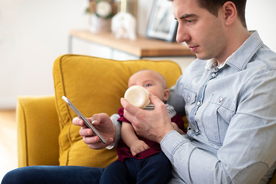 Father Checking Messages On Mobile Phone Whilst Feeding Bottle To Baby Son