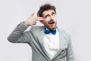 Portrait of crazy handsome bearded man in casual grey suit and blue bow tie standing with hand pistol gun gesture on his head. indoor studio shot, isolated on light grey background.