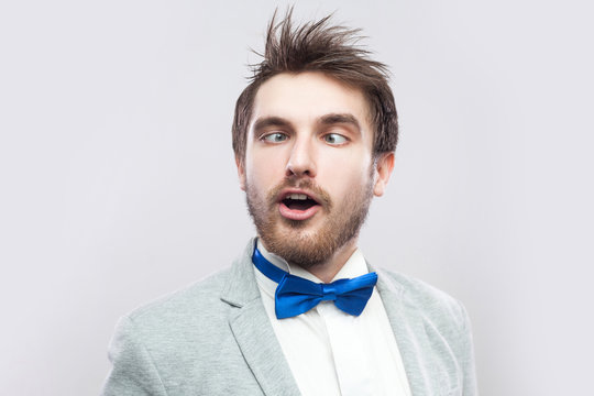 Closeup portrait of crazy funny handsome bearded man in casual grey suit and blue bow tie standing with crossed eyes and looking. indoor studio shot, isolated on light grey background.