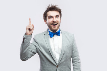 have Idea. Portrait of handsome excited bearded man in casual grey suit and blue bow tie standing and looking at camera with toothy smile. indoor studio shot, isolated on light grey background.