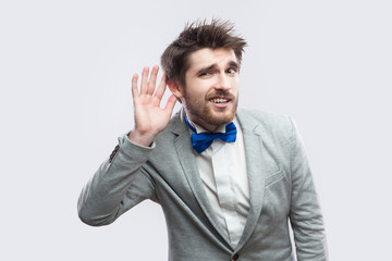 Portrait of attentive handsome bearded man in casual grey suit and blue bow tie standing holding hand near ear and trying to hear. indoor studio shot, isolated on light grey background.