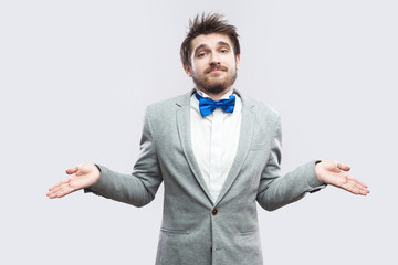 I don't know. Portrait of confused handsome bearded man in casual grey suit and blue bow tie standing with raised arms and looking at camera. indoor studio shot, isolated on light grey background.