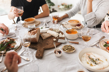 Close up group of young people sitting at the table full of delicious food having lunch together. Fresh bread with cheese on wood board in cafe