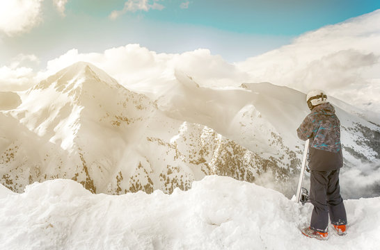 Snowboarder stay with snowboard in hands sits on big rock on mountains backdrop. Bansko, Bulgaria