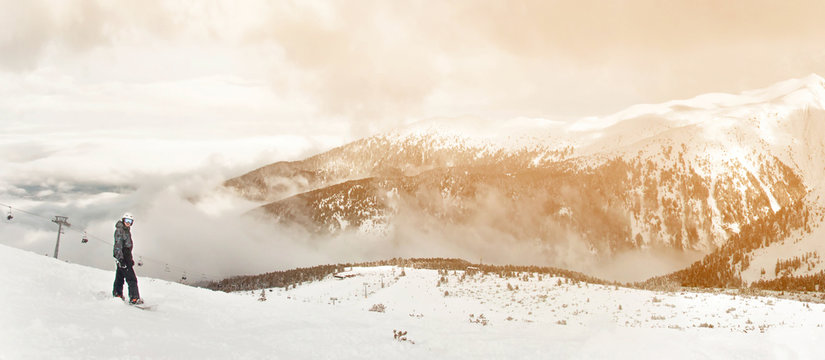 Panorama with snowboarder sit with snowboard in hands sits on big rock on mountains view. Bansko, Bulgaria