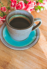 Close up view of a blue vintage cup of black coffee and a bouquet of pink carnation flowers on wooden background illuminated by sun rays coming from a side window. Good morning coffee or hello spring 