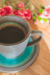 Close up view of a blue vintage cup of black coffee and a bouquet of pink carnation flowers on wooden background illuminated by sun rays coming from a side window. Good morning coffee or hello spring 