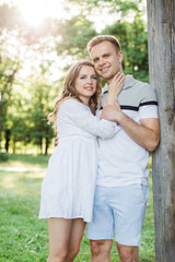 Fototapeta premium Young pretty couple in love standing in park. Handsome blonde girl in white dress hugging her boyfriend