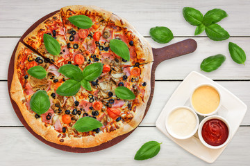 Top view of a traditional italian pizza with: ham,  mushrooms, cherry tomatoes, mozzarella, black olives decorated with basil leaves and set of sauces.