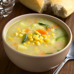 Vegetarian corn and courgette chowder served in bowl, glass of water and bread roll in the back, photographed on dark wood with natural light (Selective Focus, Focus in the middle of the soup)