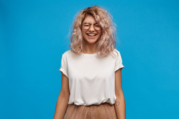 Emotional charming young European female in trendy glasses laughing, closing eyes and smiling...