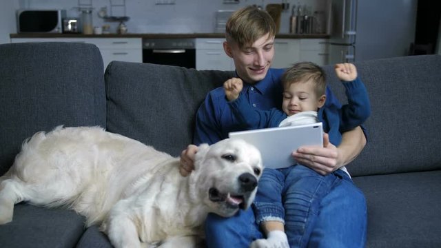 Caring young father with cheerful toddler son on his lap streaming online cartoons on digital tablet and smiling. Positive man petting labrador dog while bonding with cute little boy on sofa at home.
