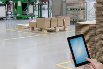 Empty production hall with folded paper boxes on wooden pallets and hands holding tablet with blank screen ready for your text.