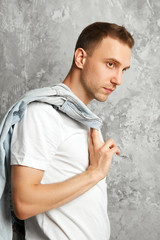 A young guy holds a denim jacket with hand standing sideways next to a concrete wall