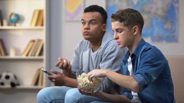 Anxious multiracial friends eating popcorn and watching TV show, leisure time