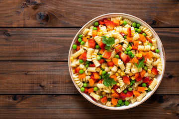 Obraz na płótnie Canvas Italian pasta salad with fusilli, corn, boiled carrot, fresh green peas and sweet pepper on wooden rustic background. Top view