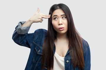 Portrait of lonely depressed beautiful brunette asian young woman in casual blue denim jacket with makeup standing with gun pistol on head. indoor studio shot, isolated on light grey background.