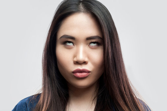 Closeup portrait of funny crazy beautiful brunette asian young woman in casual blue denim jacket with makeup standing with crossed eyes. indoor studio shot, isolated on light grey background.