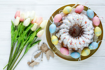 Happy Easter! Golden tray with plate wit cake and hand painted colorful eggs, tulips on white wooden table. Close up. Decoration for Easter, festive background.
