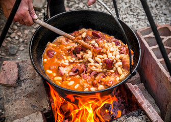 Cooking beans with bacon and sausages outdoors in black pot at picnic party on fire