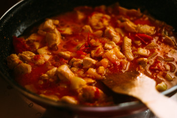 stir with a wooden spatula, cook. On the stove there is a frying pan with chicken in tomato sauce. Prepare a meal