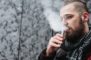 Men with beard hold and smoke his electronic cigarette outdoor in black, grey backgraund