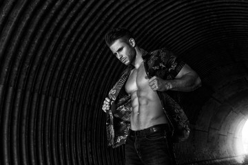 Fototapeta na wymiar Muscular young man with beard on dark tunnel urban background. Fashion portrait of brutal strong muscle guy with modern trendy hairstyle. Model, fashion concept. Sexy naked torso. Black and white.