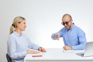 Fototapeta na wymiar Laughing brutal boss in sunglasses in a blue shirt looks at the camera with his blonde caucasian colleague sits by the desk in the office with the white background.