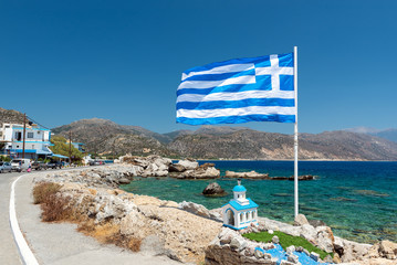 Big blue Greek flag and small church at coast of Palaiochora town on Crete island, Greece